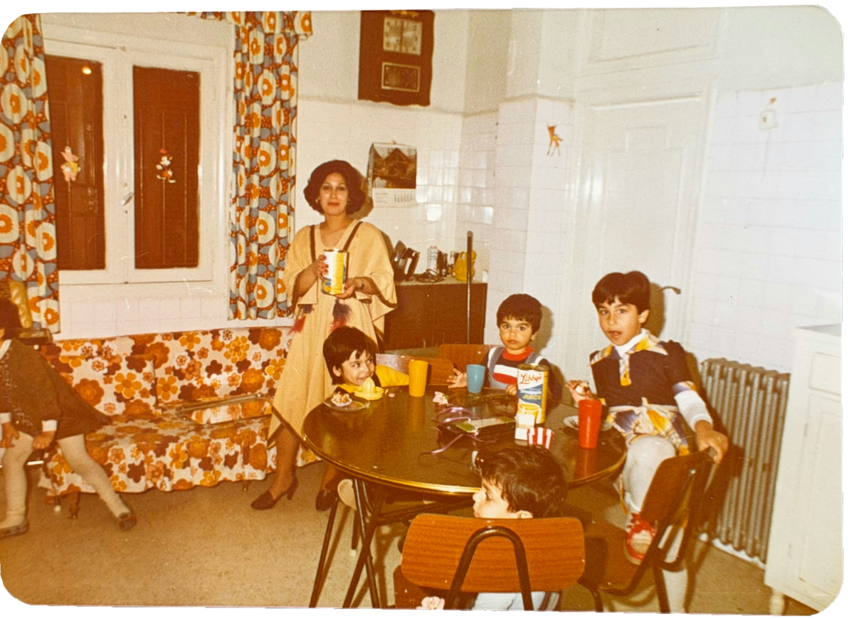 A vintage color photograph showing a family in a retro-style home interior. Teta  in a beige dress stands near a table holding a drink, while mom and siblings are seated around the table. Lebanon 1977.