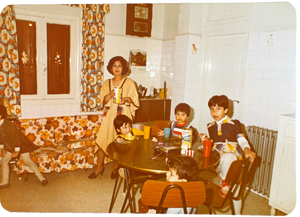 A vintage color photograph showing a family in a retro-style home interior. Teta  in a beige dress stands near a table holding a drink, while mom and siblings are seated around the table. Lebanon 1977.